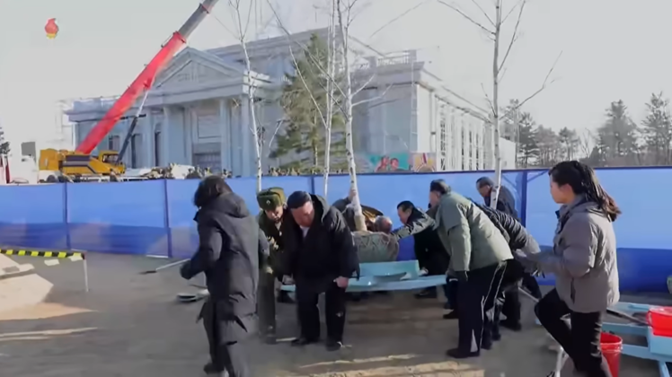 김정은이 지게를 힘껏 들어올리자 주변에 있던 간부들이 달려온다. / 화면= 조선중앙TV